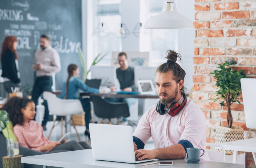 a young man working on his laptop with his co-workers working and talking behind